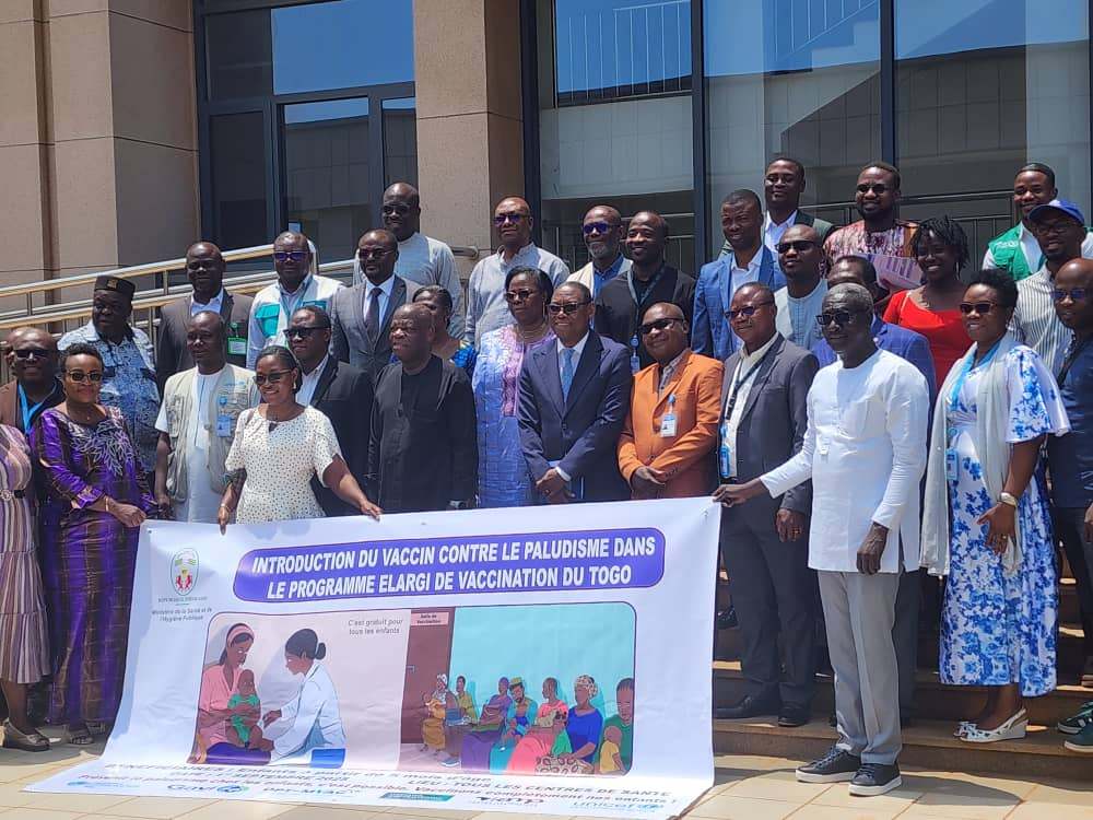 Group of people posing for a photo in front of a building, holding a banner about the introduction of the malaria vaccine in Togo’s Expanded Immunization Program