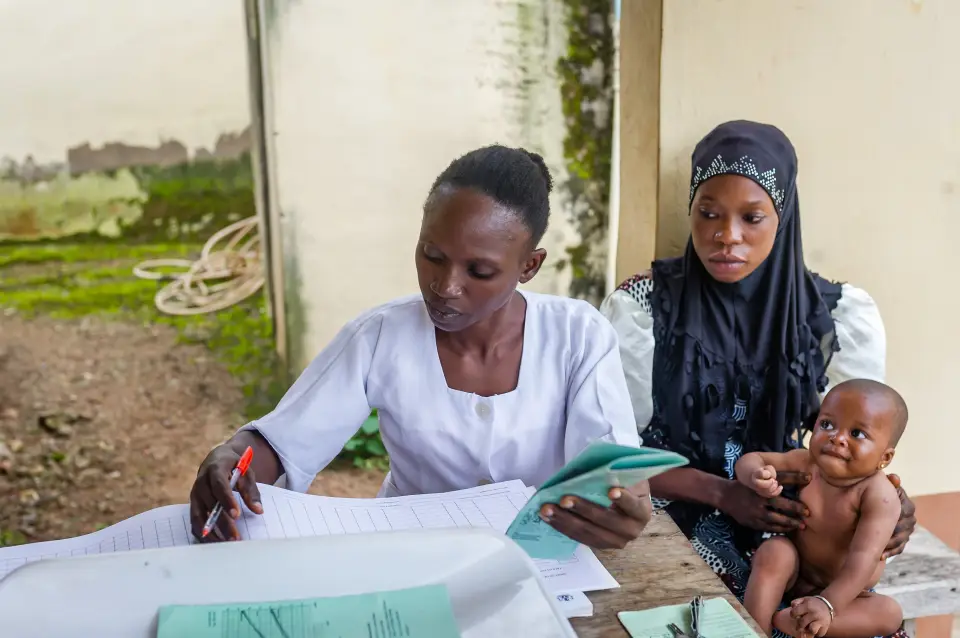 A healthcare worker in a white uniform writes in a record book while holding a patient file, sitting next to a mother dressed holding her baby on her lap.