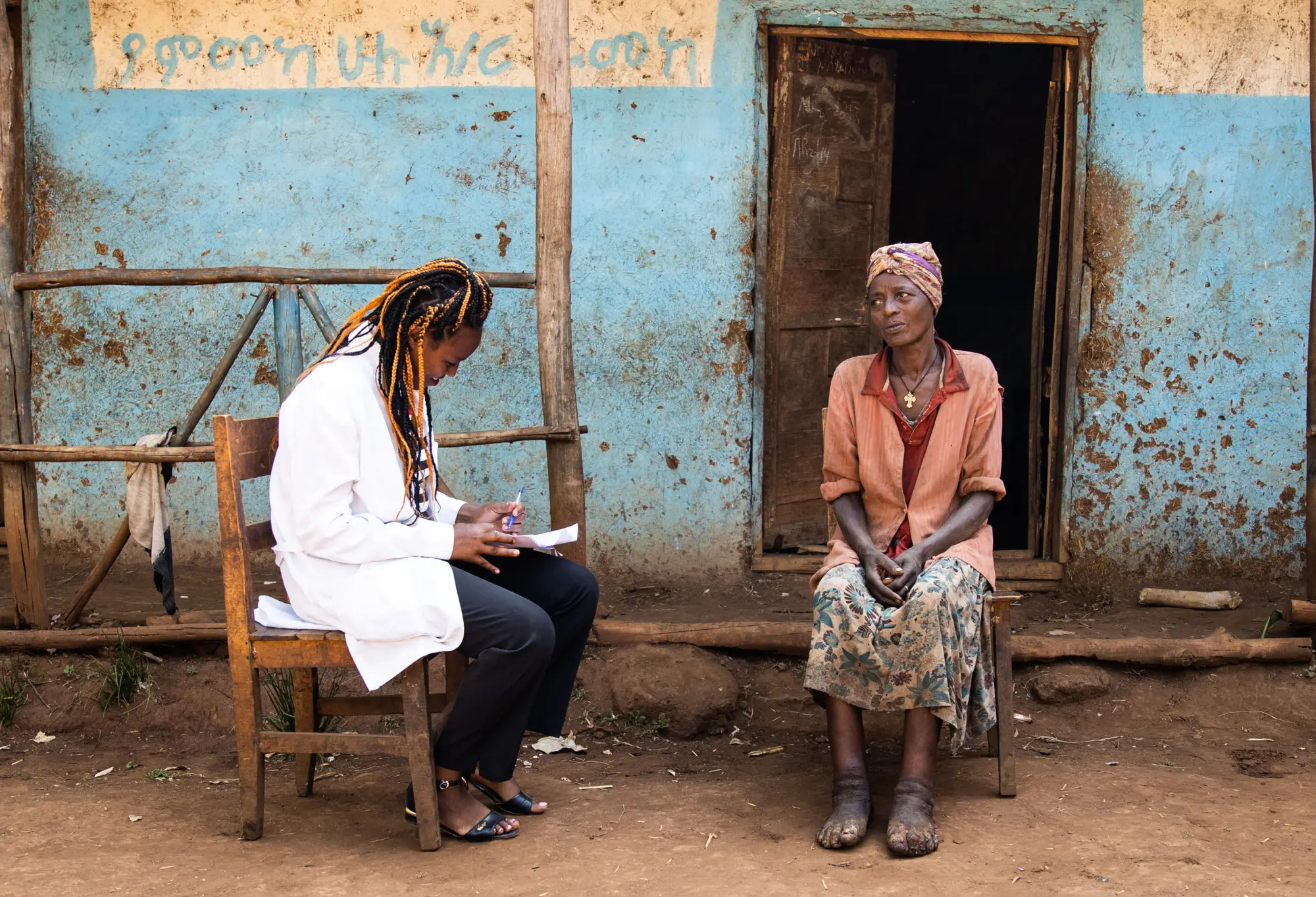 A healthcare worker wearing a white coat sits on a wooden chair, writing notes on a clipboard while speaking with an older woman.