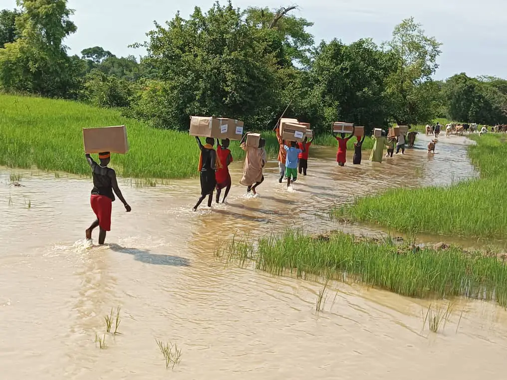 A group of women and young people volunteer to carry antimalarial medicines across a flooded road to Ayai Primary Health Care Centre in Aweil South County, South Sudan